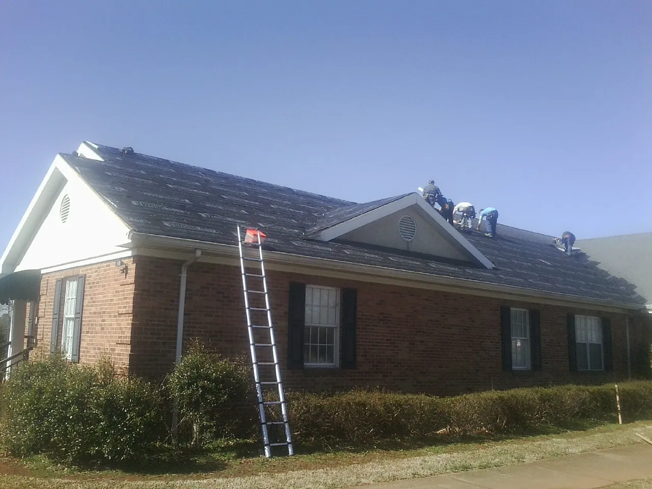 Crew tearing shingles off a brick ranch roof with a ladder set — Metal Roof Repair work by our crew in Kearsarge