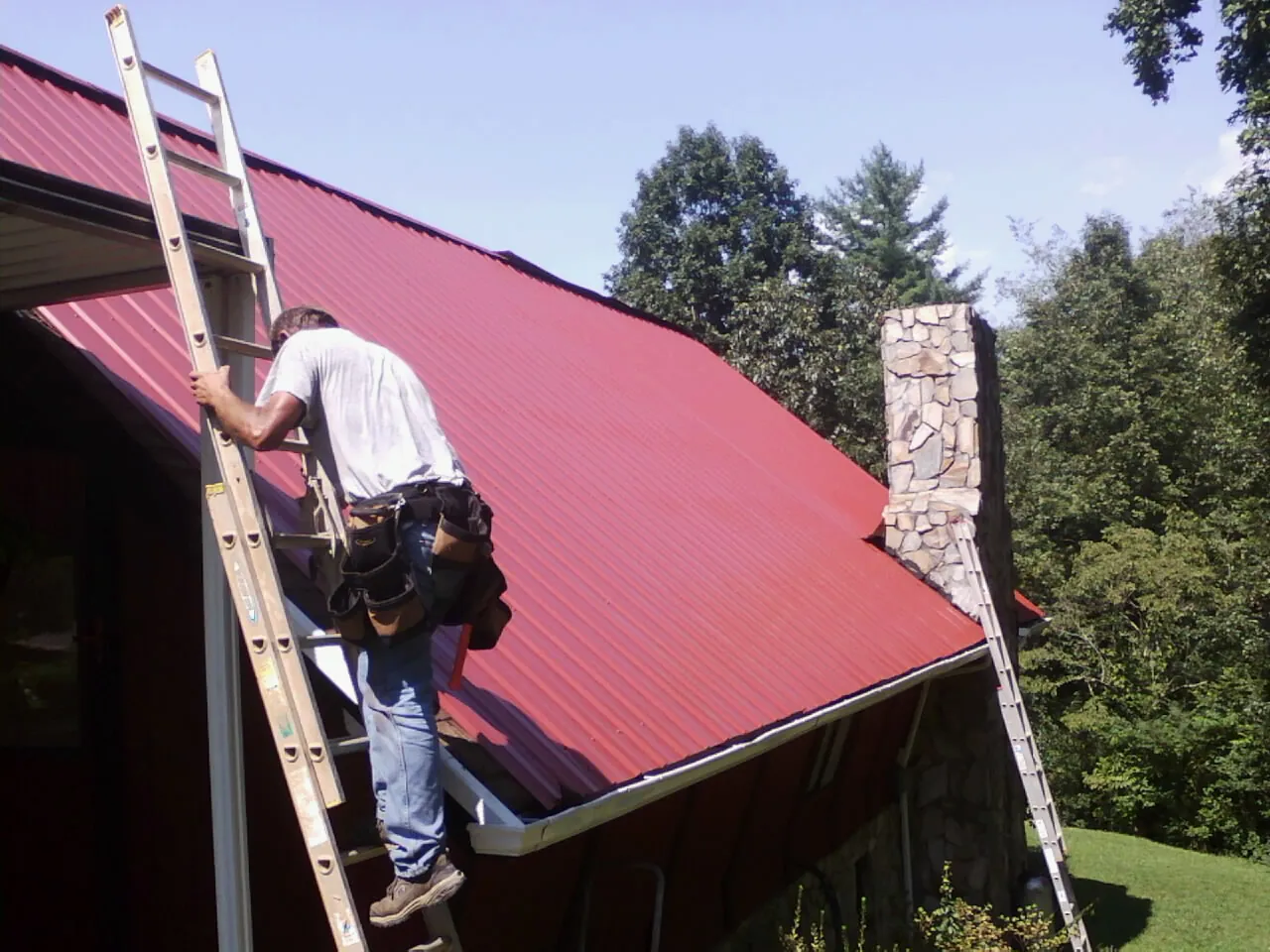 Roofer on a ladder beside a red metal roof and stone chimney — Soffit & Fascia Repair by Roofing Brothers in Kearsarge